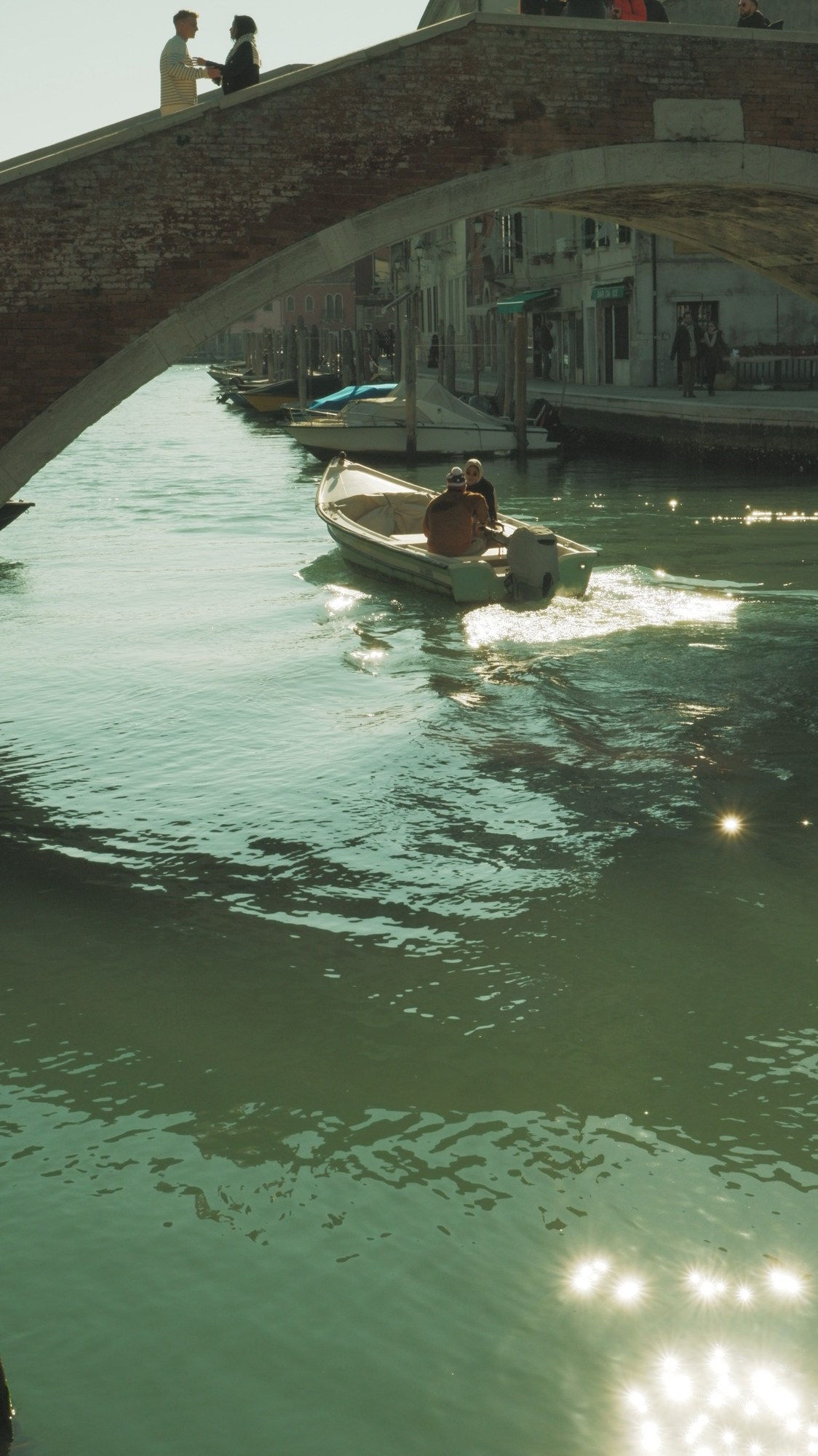 A small boat passing under a brick bridge on a sunlit canal
